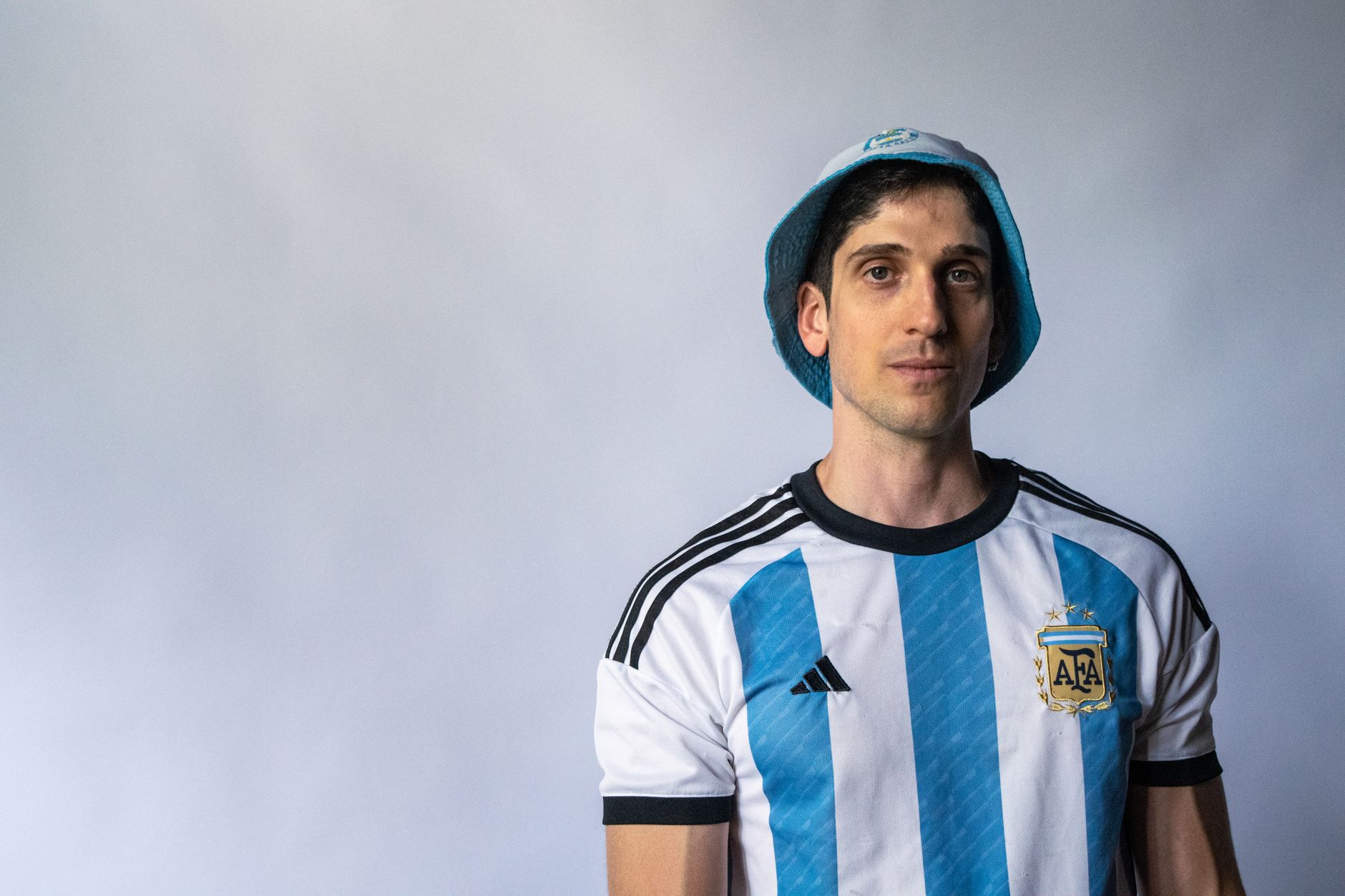 Close-up portrait of a man wearing the Argentine football jersey and hat, set against a plain backdrop.
