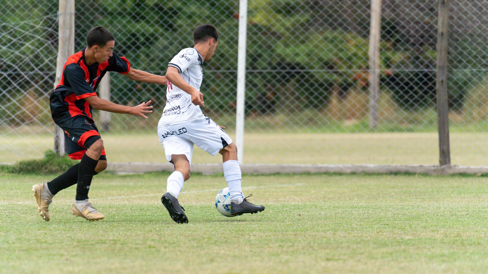 Two soccer players in action during a match on a lush green field under a mesh fence backdrop.