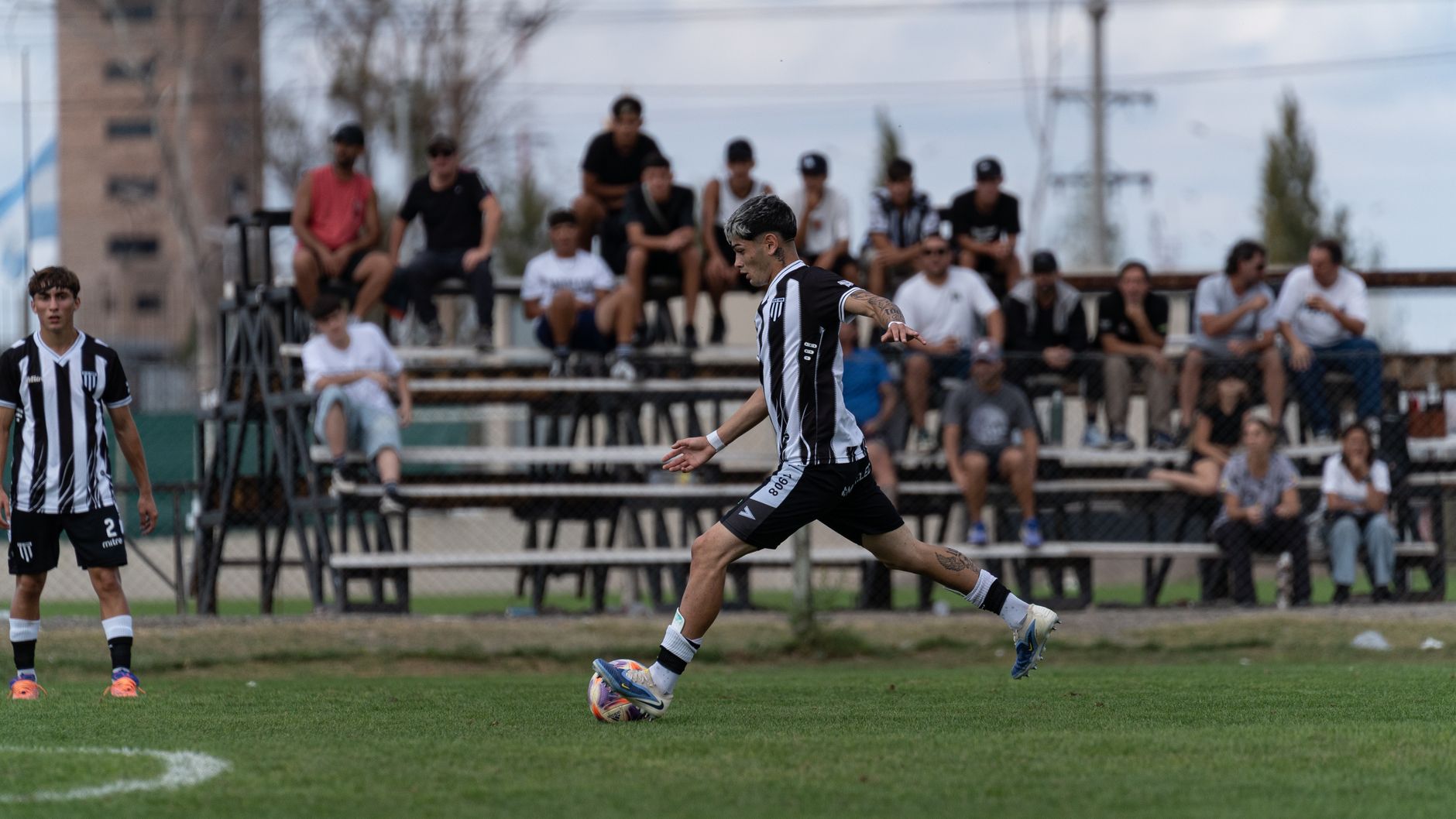 A soccer player in black and white jersey kicks the ball during a match with spectators in background.